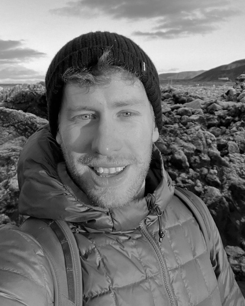 Will, wearing a dark beanie and a padded jacket, standing on a lava field at sunset.
